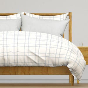 May include: A bed with a wooden frame and a white and blue plaid duvet cover. Two white pillows are visible on the bed. The bed is set against a white wall, with a wooden bedside table.