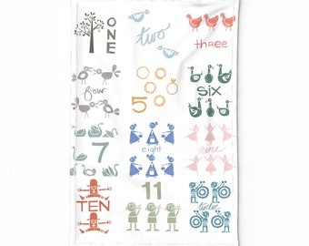 May include: White kitchen towel with colorful illustrations of the numbers one through twelve. Each number is accompanied by a corresponding image, such as birds, rings, and dancers. The towel is made of a soft, absorbent material.
