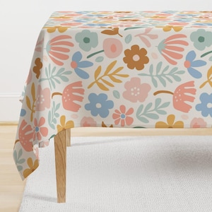 May include: A white tablecloth with a floral pattern featuring pink, blue, yellow, and orange flowers. The tablecloth is laid on a table with wooden legs.