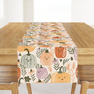May include: A beige table runner with a repeating pattern of orange, green, and purple pumpkins with smiling faces. The pumpkins are surrounded by green leaves and flowers.