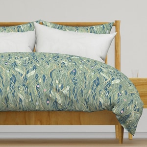 Può includere: Un letto con struttura in legno e copripiumino con design botanico. Il copripiumino presenta un motivo di forme vegetali verdi, blu e beige, con cuscini bianchi. La struttura del letto e il comodino sono in legno chiaro.