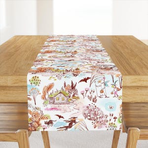 May include: A white table runner with a whimsical Halloween design featuring a cottage, bats, a witch, a cat, and mushrooms. The design is in shades of brown, orange, and green.