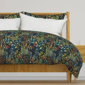 Puede incluir: Una cama con una funda nórdica y fundas de almohada azul marino. La funda nórdica presenta un colorido estampado botánico con ilustraciones de plantas verdes, amarillas, naranjas y rojas. Las almohadas blancas contrastan con el fondo oscuro. La estructura de la cama es de madera clara.