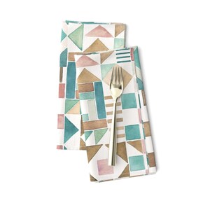 May include: Two white fabric napkins with a geometric pattern in shades of brown, teal, and pink. The napkins are folded and a gold fork is placed on top.
