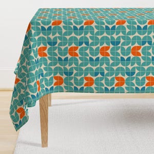 Pode incluir: Uma toalha de mesa retangular com um padrão geométrico retrô. O design apresenta formas nas cores verde-azulado, laranja e creme dispostas em um padrão repetitivo. A toalha de mesa está sobre uma mesa de madeira, mostrando seu design.