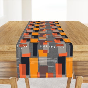 May include: A gray, orange, and black geometric patterned table runner. The runner has a modern abstract design with rectangular shapes and white dots.