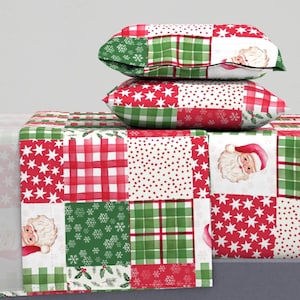 May include: Christmas-themed patchwork pillows and table runner. The design features red and green squares with stars, snowflakes, plaid, and Santa Claus illustrations. The pillows are stacked on top of the table runner, creating a festive display.
