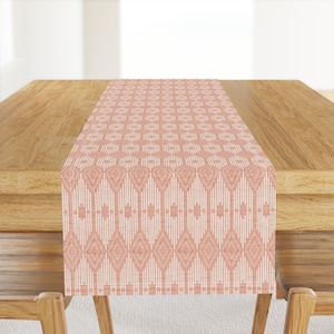 Op de afbeelding: Een roze en witte geometrische tafelrunner. De runner ligt op een houten tafel.