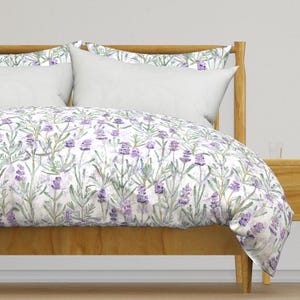 May include: A bed with a wooden frame and a white duvet cover and pillow set. The duvet cover features a lavender floral pattern with purple flowers and green leaves on a white background. The pillows are plain white.