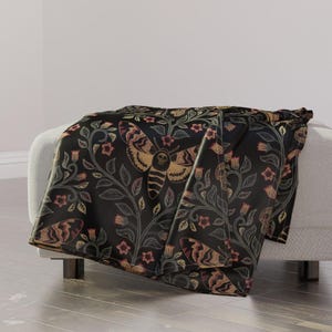 May include: Black throw blanket featuring a repeating pattern of a skull-faced moth, flowers, and vines. The moth is a light brown colour with black markings and a skull on its chest. The flowers are red and the vines are green.
