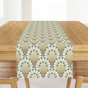 May include: A white table runner with a repeating pattern of blue and gold stylized lotus flowers. The runner is on a wooden table.