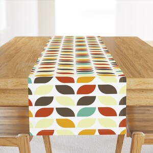 May include: A white table runner with a geometric pattern of brown, orange, yellow, and teal leaves. The runner is on a wooden table.