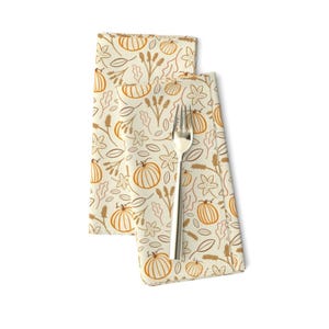 May include: Two cream-colored cloth napkins with a repeating pattern of orange pumpkins, leaves, and floral elements. A silver fork rests on the napkins. The napkins are folded and displayed on a white background.