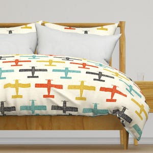 May include: A white duvet cover with a repeating pattern of colorful airplanes. The airplanes are in shades of yellow, red, blue, and black. The duvet cover is on a wooden bed frame with a white pillow and a yellow pillow.