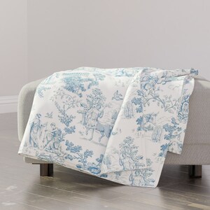 May include: A white throw blanket with a blue toile pattern featuring a repeating design of trees, birds, and figures. The blanket is folded and draped over a light gray ottoman.