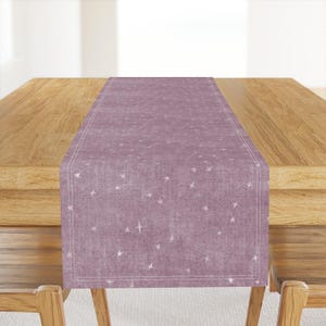 May include: A dusty rose table runner with a white star pattern. The rectangular runner is centered on a wooden table, with a white stitched border. The stars are evenly spaced across the fabric.