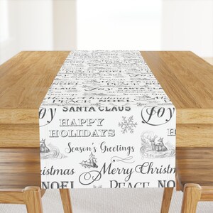 May include: White table runner with a black and gray print of various Christmas greetings, including "Happy Holidays", "Season's Greetings", "Merry Christmas", and "Peace Noel".