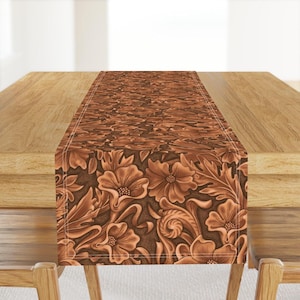 May include: A brown table runner with a floral pattern. The pattern is embossed and looks like leather. The runner is on a wooden table.
