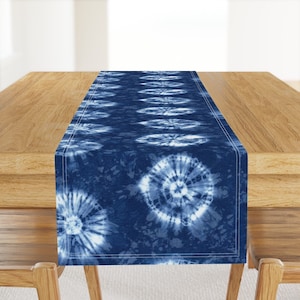 May include: A blue and white tie-dye table runner with a circular pattern. The runner is on a wooden table.