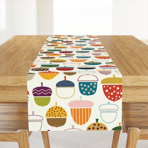 May include: A white table runner with a pattern of colorful acorns. The acorns are in various shapes and sizes, with different patterns and colors. The runner is perfect for adding a touch of fall decor to your table.