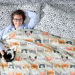 May include: A woman wearing glasses lies in bed with a black and white cat. The bedding features a pattern of cats wearing glasses.