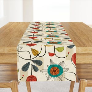 May include: A white table runner with a colorful floral pattern. The pattern features stylized flowers with red, yellow, green, blue, and orange accents. The runner is perfect for adding a touch of color and whimsy to any dining table.