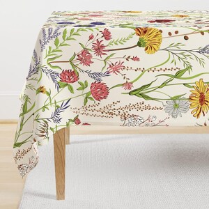 May include: A rectangular tablecloth featuring a cream-colored background with a botanical print of various wildflowers in shades of yellow, pink, red, purple, and green. The pattern includes detailed illustrations of daisies, clover, and grasses.