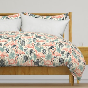 Puede incluir: Una funda nórdica blanca con un estampado de flamencos rosas y hojas tropicales. La funda nórdica está sobre un marco de cama de madera con dos almohadas blancas.