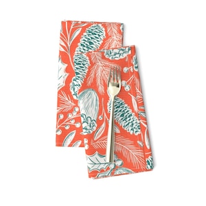 May include: Two orange napkins with a white and teal pine cone and holly design. The napkins are folded and a silver fork is placed on top.