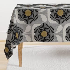 May include: A gray and white tablecloth with a repeating pattern of black and gold stylized flowers. The tablecloth is draped over a table with wooden legs.