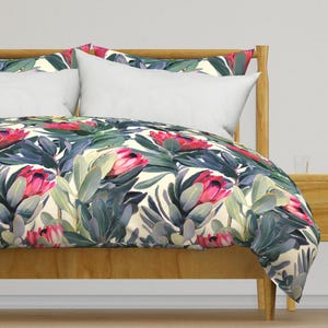 May include: A bed with a wooden frame and white pillows. The bedding features a floral pattern with large pink protea flowers, green leaves, and a cream background. The overall theme is a botanical or floral design.