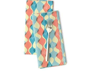 May include: Two folded napkins with a retro pattern of wavy lines in blue, yellow, and orange with white snowflake-like designs. The napkins are folded and placed on a white surface with a silver fork on top.