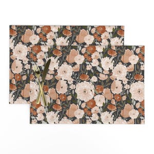 May include: Two floral placemats with a dark background. The placemats feature a repeating pattern of watercolor-style flowers in shades of peach, cream, and rust. A gold fork and knife are placed on the placemats.