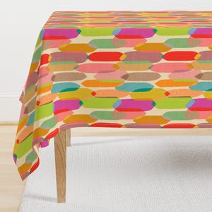 Könnte beinhalten: Eine rechteckige Tischdecke mit einem sich wiederholenden geometrischen Muster aus länglichen, abgerundeten Formen in leuchtenden Farben wie Rot, Orange, Gelb, Grün, Pink und Türkis auf einem hellen cremefarbenen Hintergrund, über einen Holztisch drapiert.