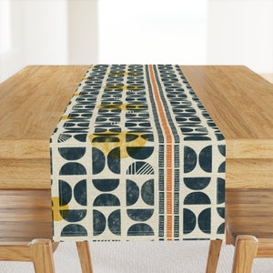 May include: A beige table runner with a geometric pattern of navy blue half circles and yellow circles. The pattern is repeated across the runner and has a vertical orange stripe down the center.