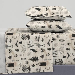 May include: Two rectangular pillows and a matching bed runner featuring a beige fabric base with a repeating black ink-style print. The pattern includes illustrations of skulls, crows, pine trees, glass bottles, candles, and various botanical elements.