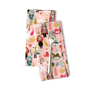 May include: Pink fabric napkins with a repeating pattern of a Nutcracker theme. The pattern includes a Nutcracker soldier, a ballerina, a Christmas tree, and other holiday elements.
