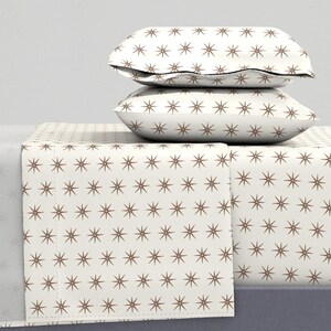 May include: White bedding with a brown star pattern. The bedding is folded and stacked on a bed. There are two white pillows with a brown star pattern on top of the bedding.