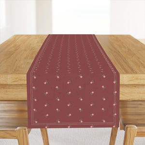Puede incluir: Camino de mesa burdeos con un patrón repetido de pequeñas flores rosas y hojas verdes. El camino de mesa tiene un borde blanco y está colocado sobre una mesa de madera. El diseño floral añade elegancia.