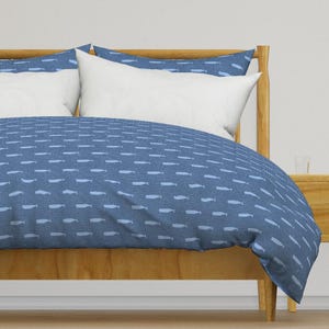 Könnte beinhalten: Blau-weißes Bettwäsche-Set mit Wal-Muster auf einem Holzrahmen. Die Bettwäsche zeigt ein sich wiederholendes Muster aus hellblauen Walen auf einem dunkleren blauen Hintergrund. Weiße Kissen ergänzen das nautische Thema.