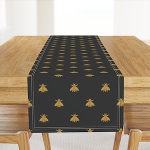May include: A black table runner with a repeating pattern of gold bee silhouettes. The runner has a white border.