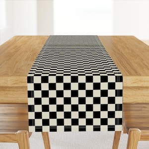 May include: A rectangular table runner featuring a repeating black and white checkered pattern. The fabric has a clean, straight-stitched border along the edges, creating a uniform grid design that spans the entire length of the textile piece.