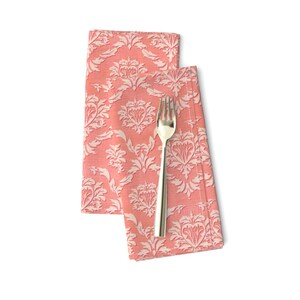 May include: Two coral pink fabric napkins with a white damask pattern. The napkins are folded and a silver fork is placed on top of them.