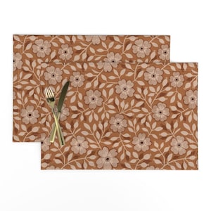 May include: Two brown placemats with a white floral pattern. The placemats have a repeating pattern of white flowers and leaves on a brown background.