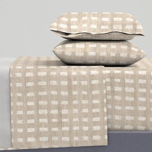 Puede incluir: Juego de cama con estampado beige y blanco. Dos almohadas apiladas sobre una sábana doblada. Las almohadas y la sábana tienen un patrón rectangular repetitivo. La ropa de cama es de color neutro y parece estar hecha de lino o algodón.