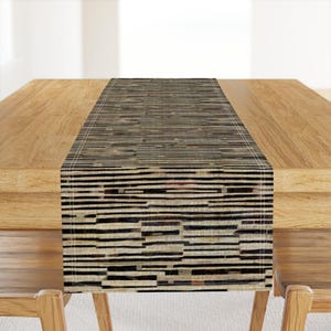 May include: A rectangular table runner featuring a horizontal pattern of thin, irregular black and dark brown stripes layered over a light beige background. The fabric has a visible woven texture with clean, straight stitched edges along the sides.