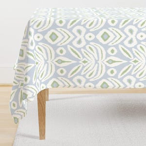 May include: A rectangular tablecloth featuring a repeating ikat-style pattern in soft sage green, white, and light blue. The fabric drapes over a table with light wooden legs, displaying a symmetrical, organic motif across the entire surface.