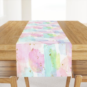 May include: A pastel pink, blue, and green watercolor table runner with gold glitter stars. The runner is on a wooden table.