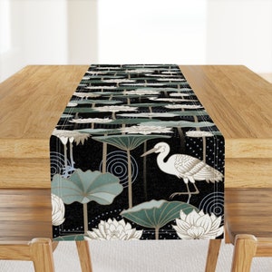 May include: A black table runner with a white egret standing on a green lotus leaf. The background is black with white dots and blue circles. The runner features a repeating pattern of lotus leaves and flowers.