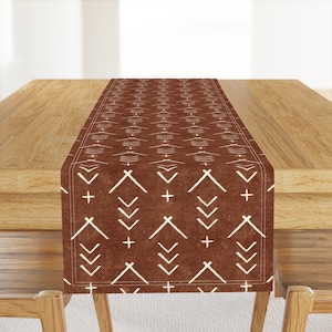 May include: A brown table runner with a white geometric pattern of arrows, crosses, and diamonds. The runner is on a wooden table.